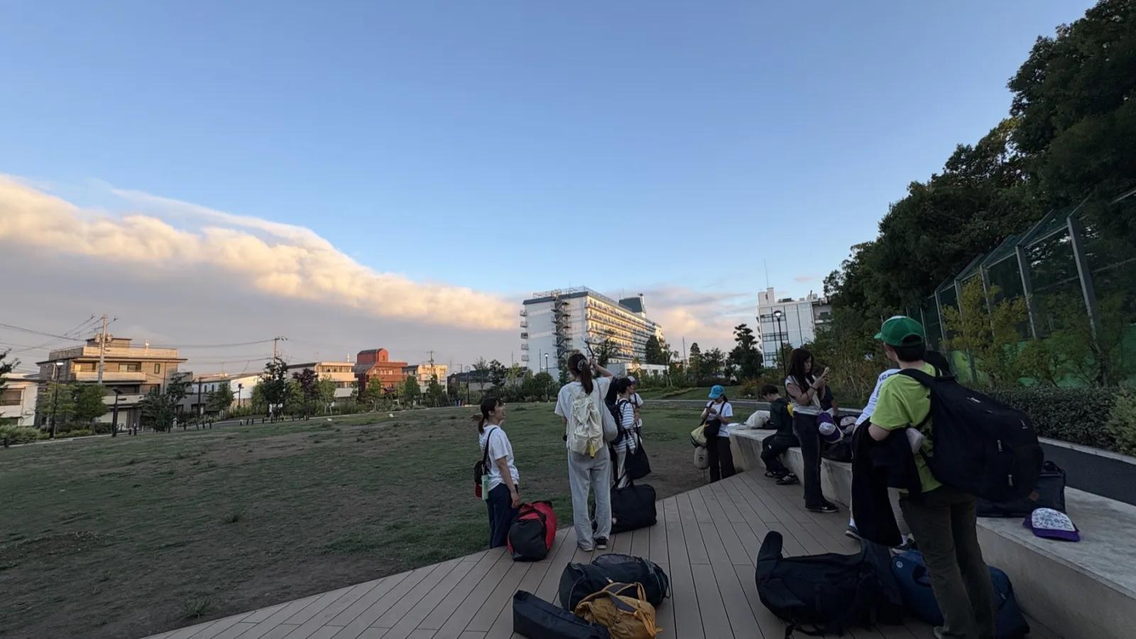 夕暮れの公園のデッキ。大きな荷物やバッグが床に並び、参加者とスタッフが数人立っている。空には夕焼けに染まった雲