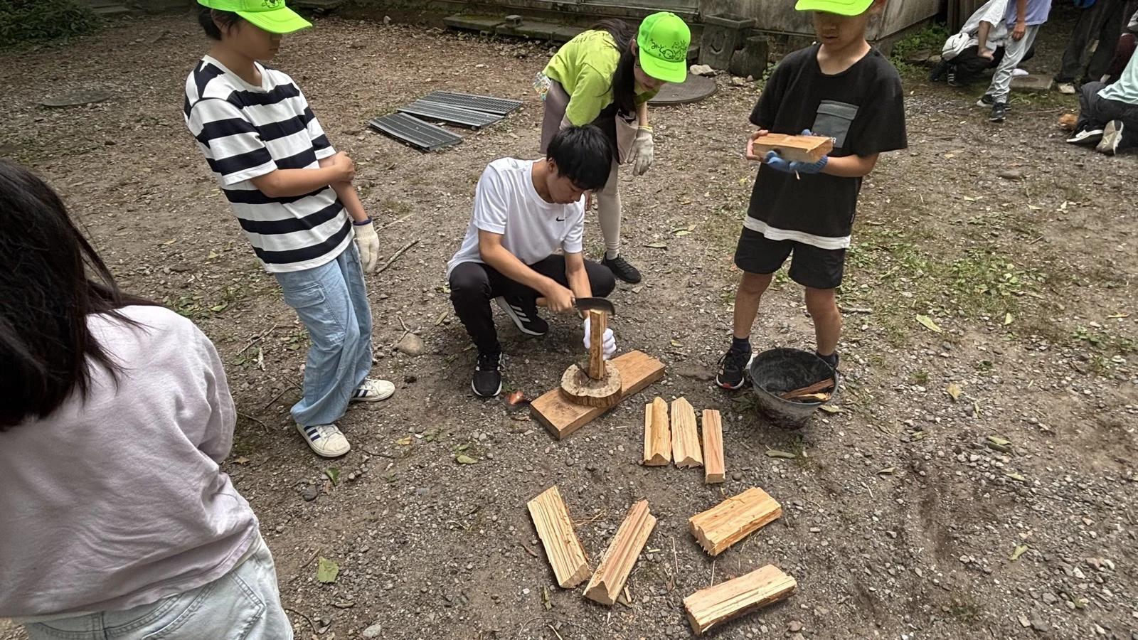 炊事場の近くで、スタッフが木片を台に立てて細く割ってみせ、参加者が手順を見ている。割った薪が地面に並んでいる