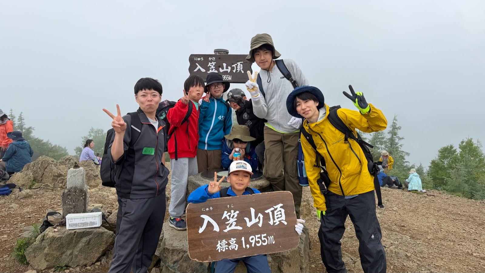 山頂の標識の前で、別のグループが「入笠山頂 標高1,955m」の看板を持って記念撮影をしている
