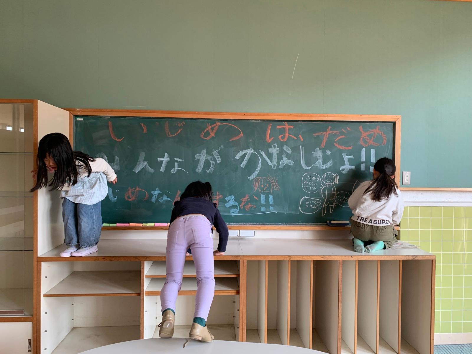 教室の黒板いっぱいに、白と色のチョークで「いじめは だめ」「みんながかなしむよ!!」「いじめなんかするな!!」と大きく書かれている。子どもたちが黒板の前に立っている