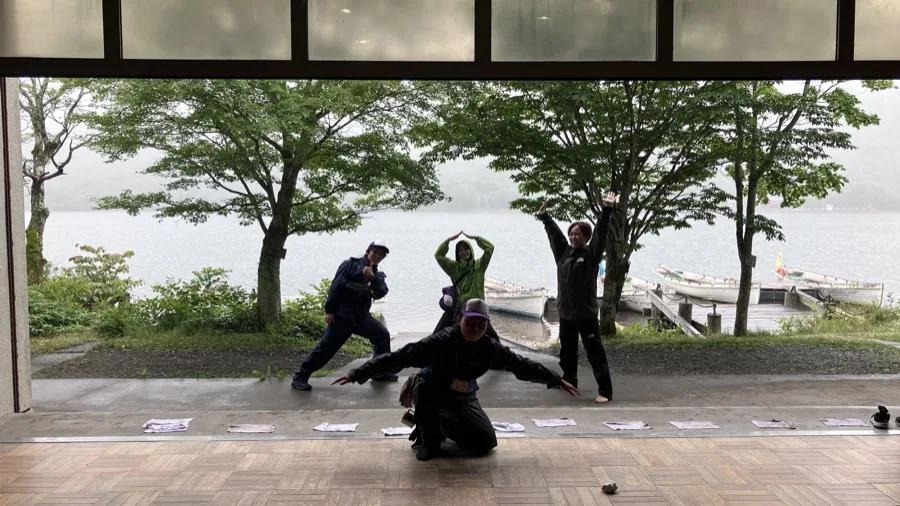 施設の広間から、大きく開いた窓越しに大沼が見える。手前のテラスで参加者4人が思い思いのポーズを取っている。湖には手こぎボートが並ぶ