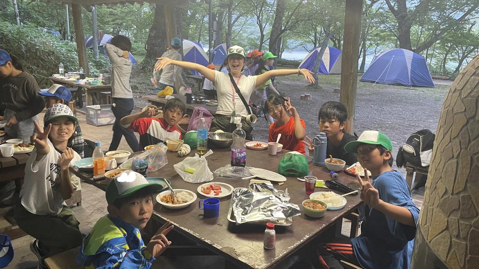 屋根付きの野外炊事場で、班のテーブルを囲んでカレーライスなどの夕食を食べながらポーズを取る参加者たち。奥にはテントと湖が見える