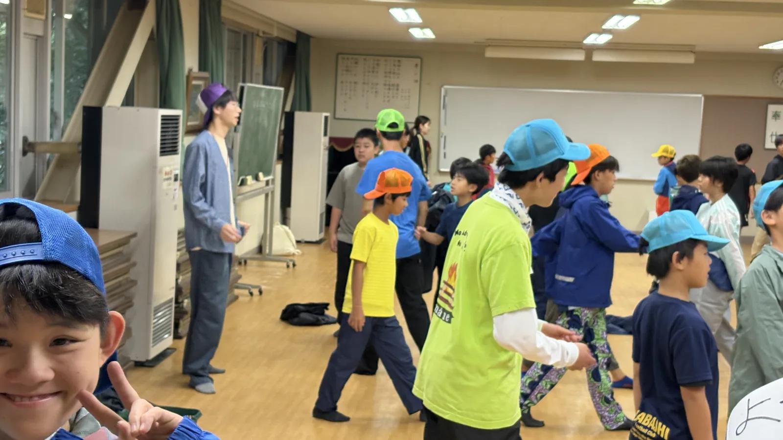 同じ広間で、参加者とスタッフが立ち上がって体を動かしている。手前の参加者がカメラに向かってピースをしている