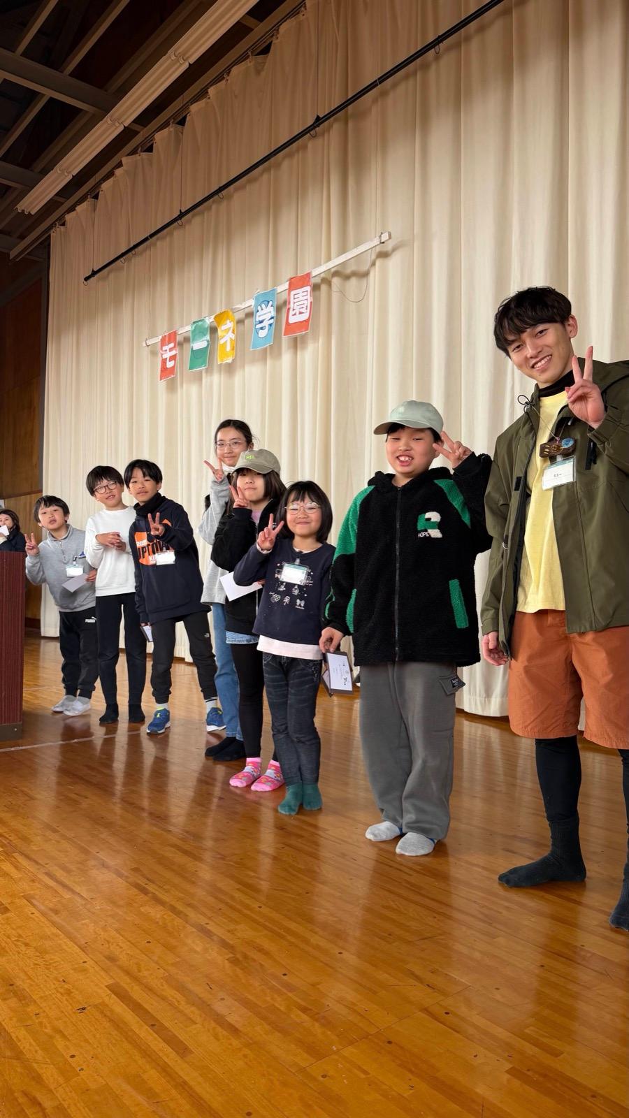 体育館の舞台に子どもたちが横一列に並び、卒業証書を手に持ってポーズをとっている。リーダーが右端に立つ。幕には「モーネ学園」の文字