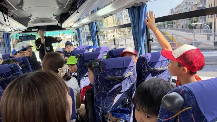バス車内の前方でマイクを持って話すスタッフ。座席から手を挙げている参加者がいる