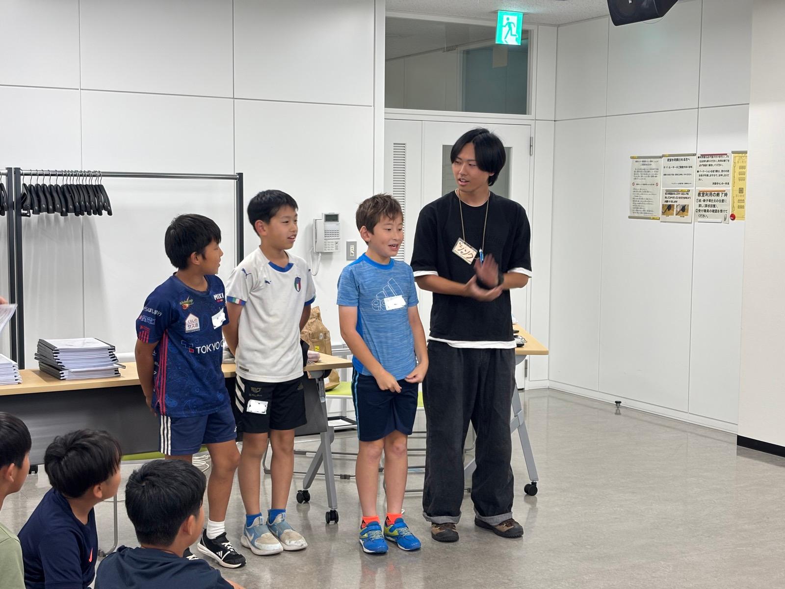 会議室で、3人の子どもとスタッフが前に立ってレクに挑戦中。奥の机には文集の束が積まれている
