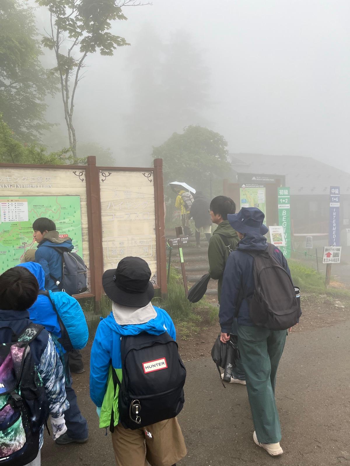 登山口付近。濃い霧のなか、レインウェアを着た子どもたちが集まっている。「入笠山ハイキングコース入口」の看板と、湿原・山頂を示す道標が立っている