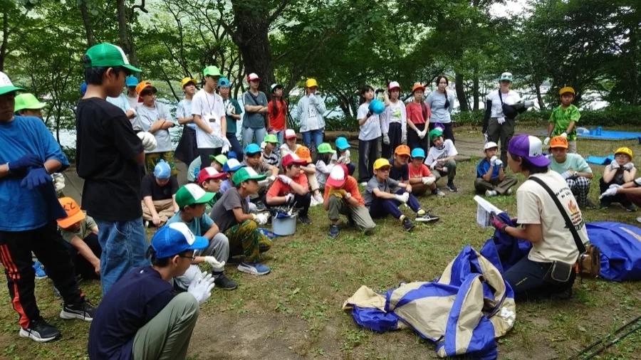 湖畔の林で、たたまれたテントを前にスタッフの説明を聞く参加者たち。全員が軍手をつけて座っている