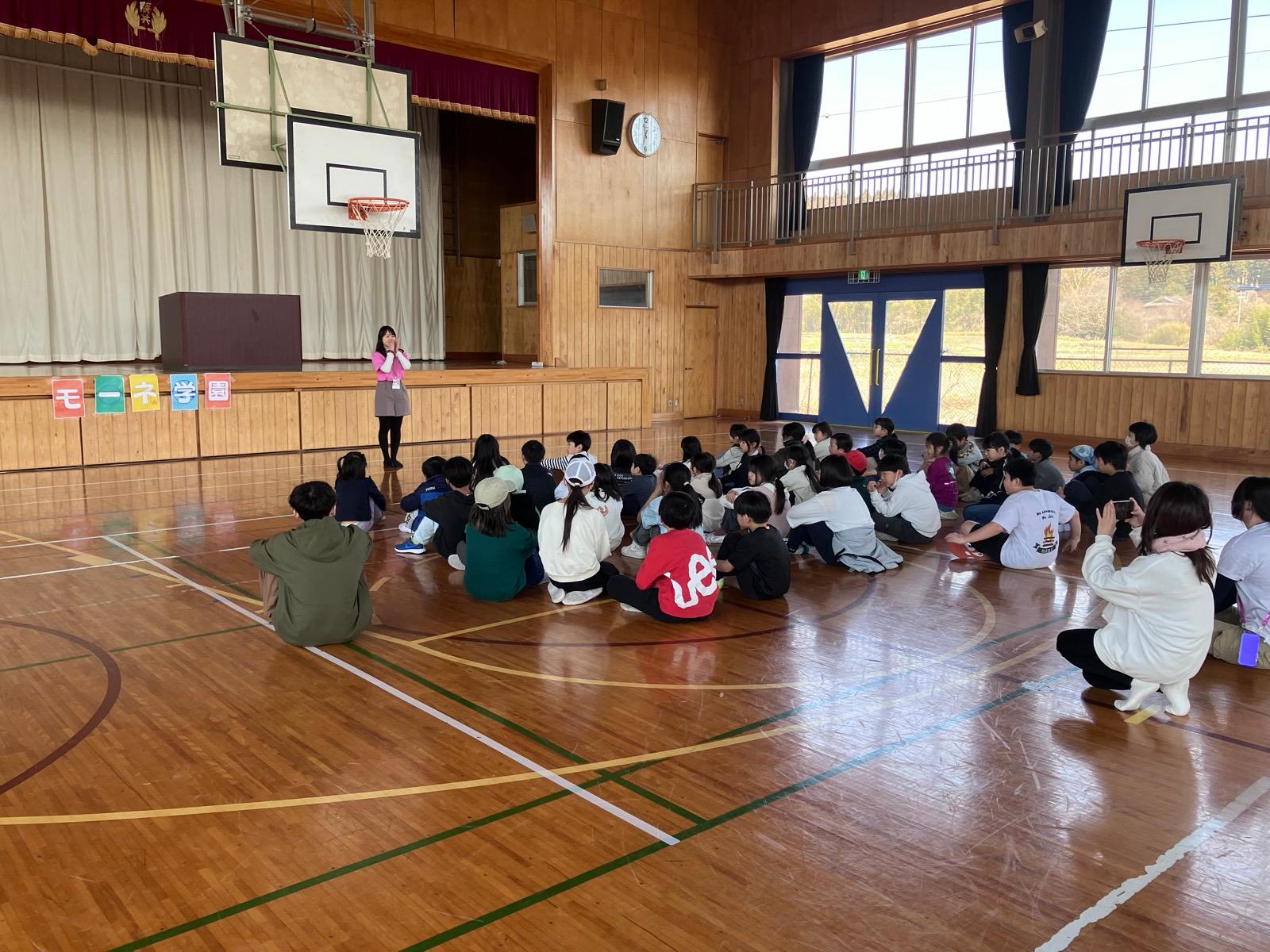 体育館の舞台の前に「モーネ学園」と色画用紙で作った文字が貼られ、その前でリーダーがマイクを持って話している。床には大勢の子どもたちが座っている