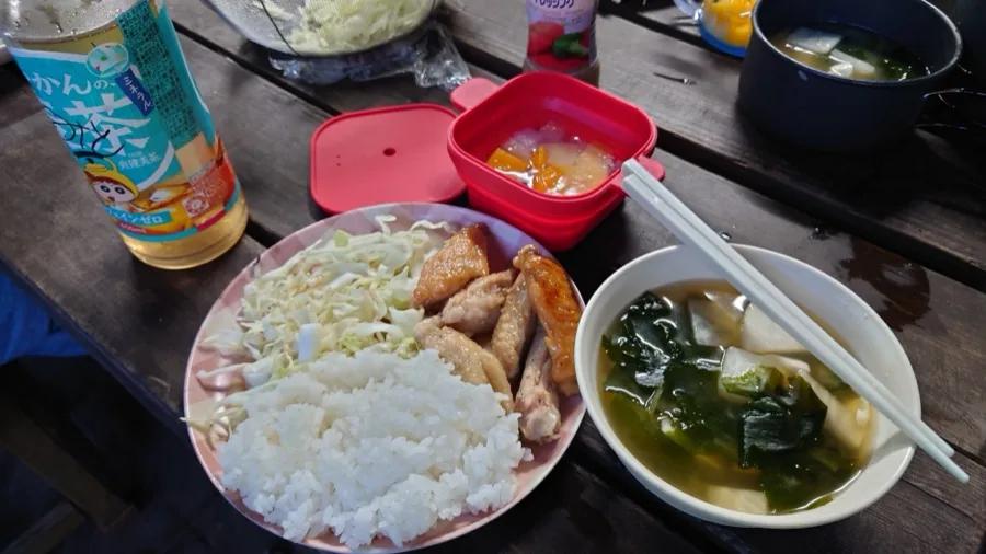 屋外の木のテーブルに並んだ夕食。ごはん、千切りキャベツ、手羽先、わかめと豆腐のみそ汁、デザートのフルーツ、ペットボトルのお茶