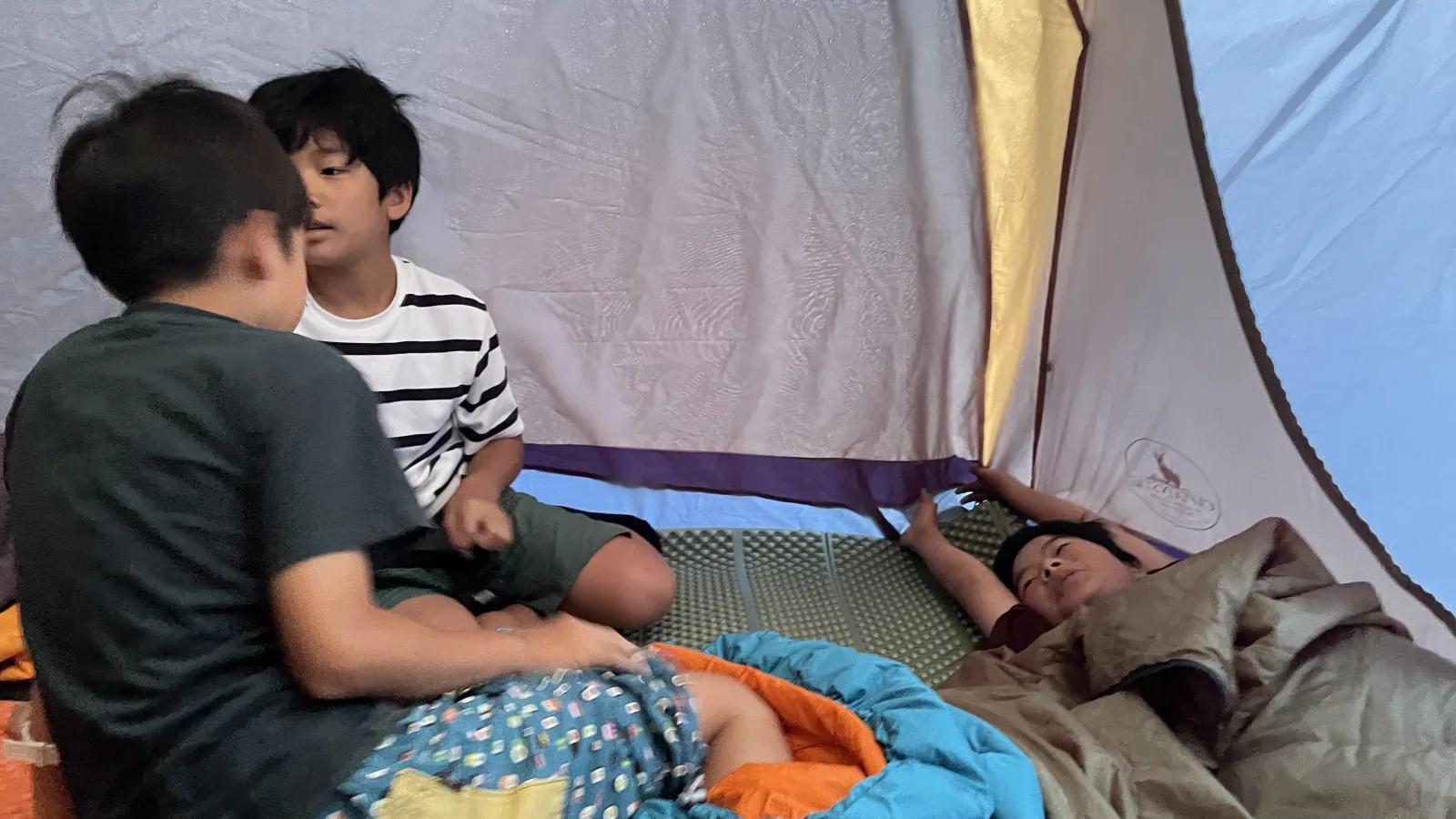 テントの中。寝袋やマットの上で、起き出した参加者たちが向かい合って話している。ひとりはまだ横になっている