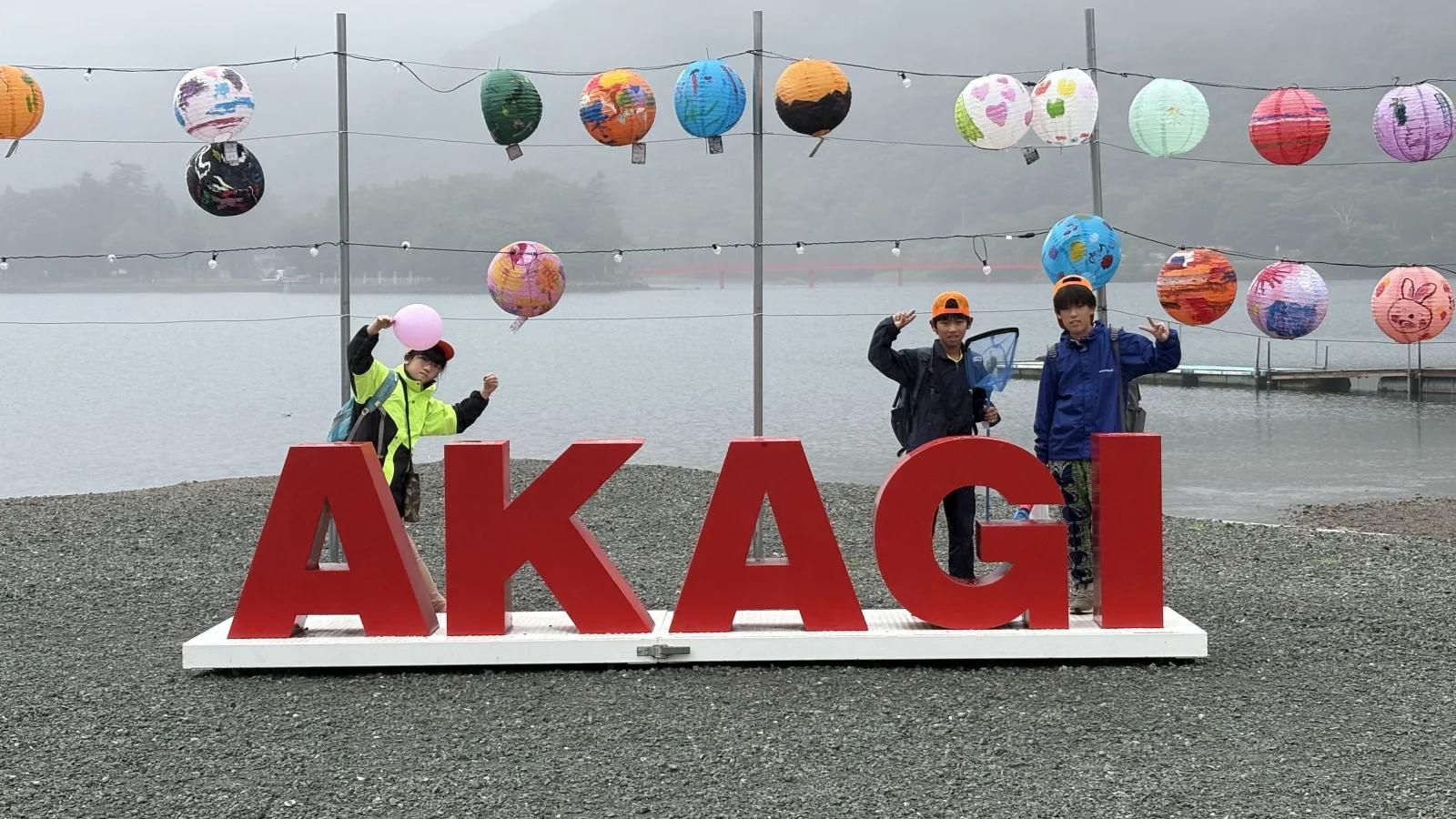 霧のかかった湖のほとり。赤い立体文字の「AKAGI」のモニュメント前で、参加者3人がポーズを取っている。上空にはカラフルな球形の飾りが吊るされている