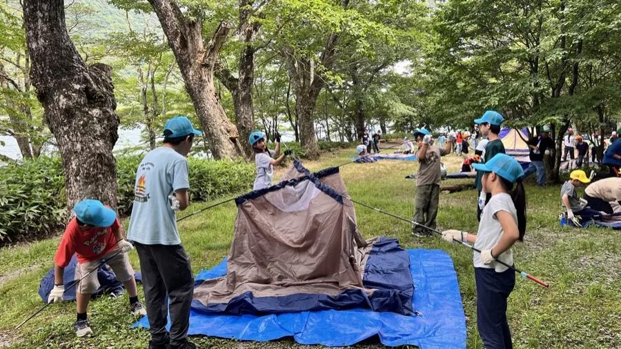 ブルーシートの上にドームテントを広げ、ポールを通して組み立てる参加者とスタッフ。周囲でも別の班が設営している