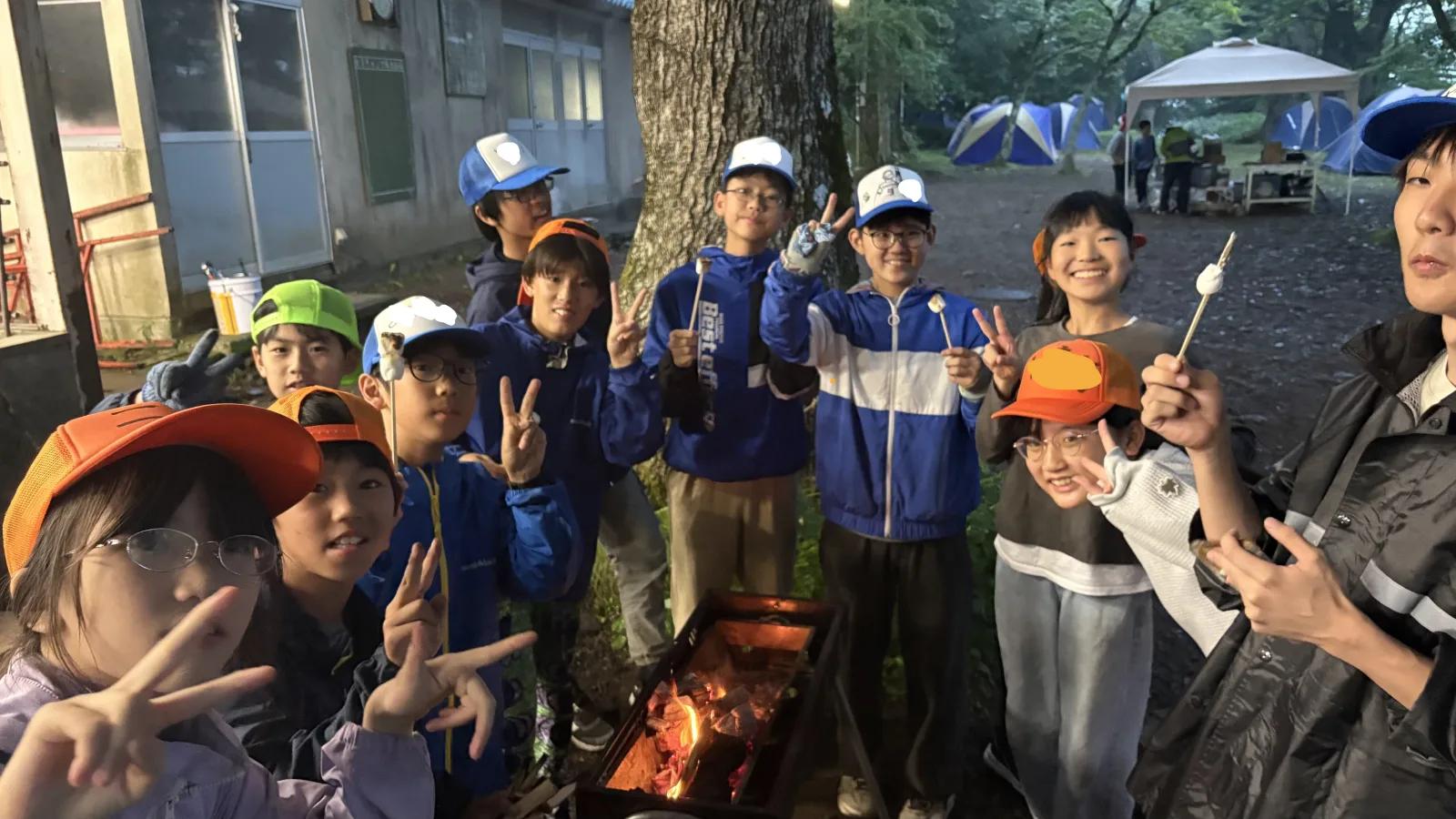 夕暮れの炊事場前。串に刺したマシュマロを手に持った参加者たちが集まって笑い、多くがピースをしている。奥にはテントとタープが見える