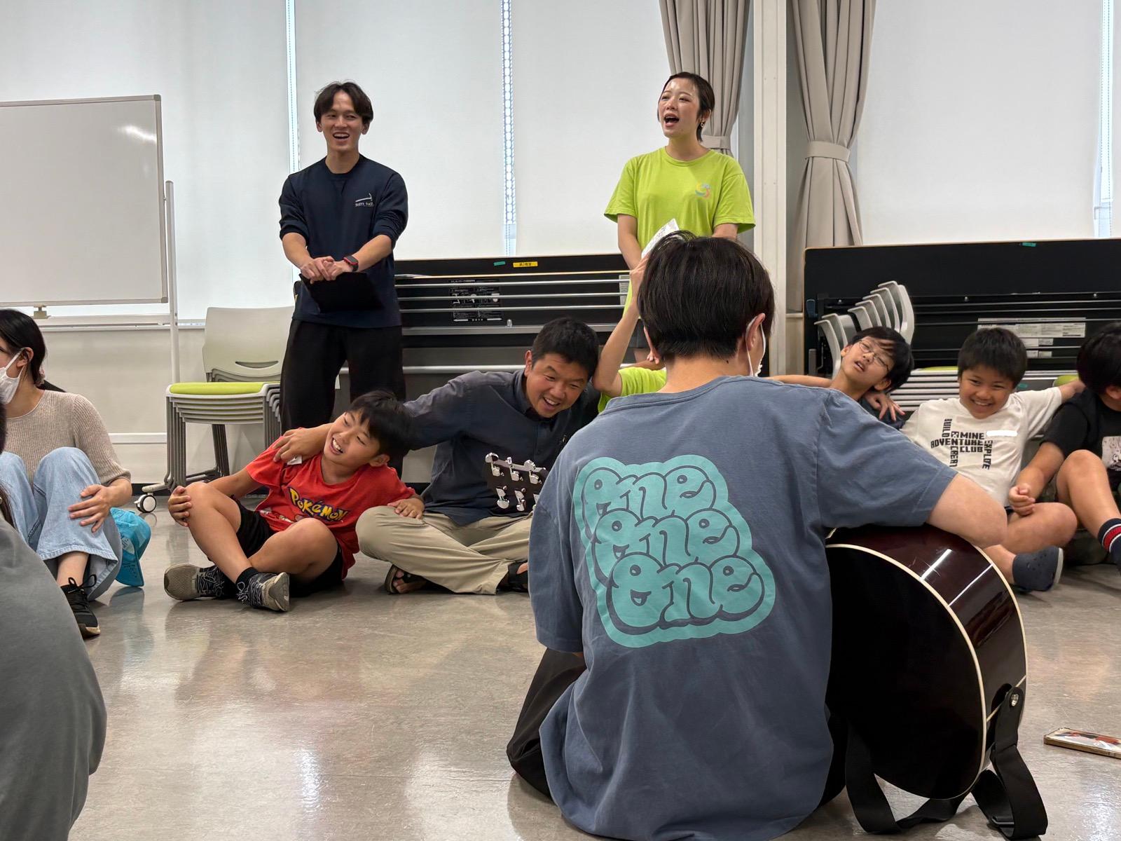 ギターを弾く人を囲んで、子どもたちとスタッフが座って歌っている。全員笑っている