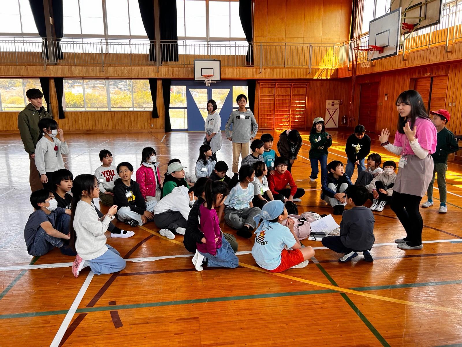 廃校の木造の体育館。床に座った子どもたちの前で、リーダーが立って話している