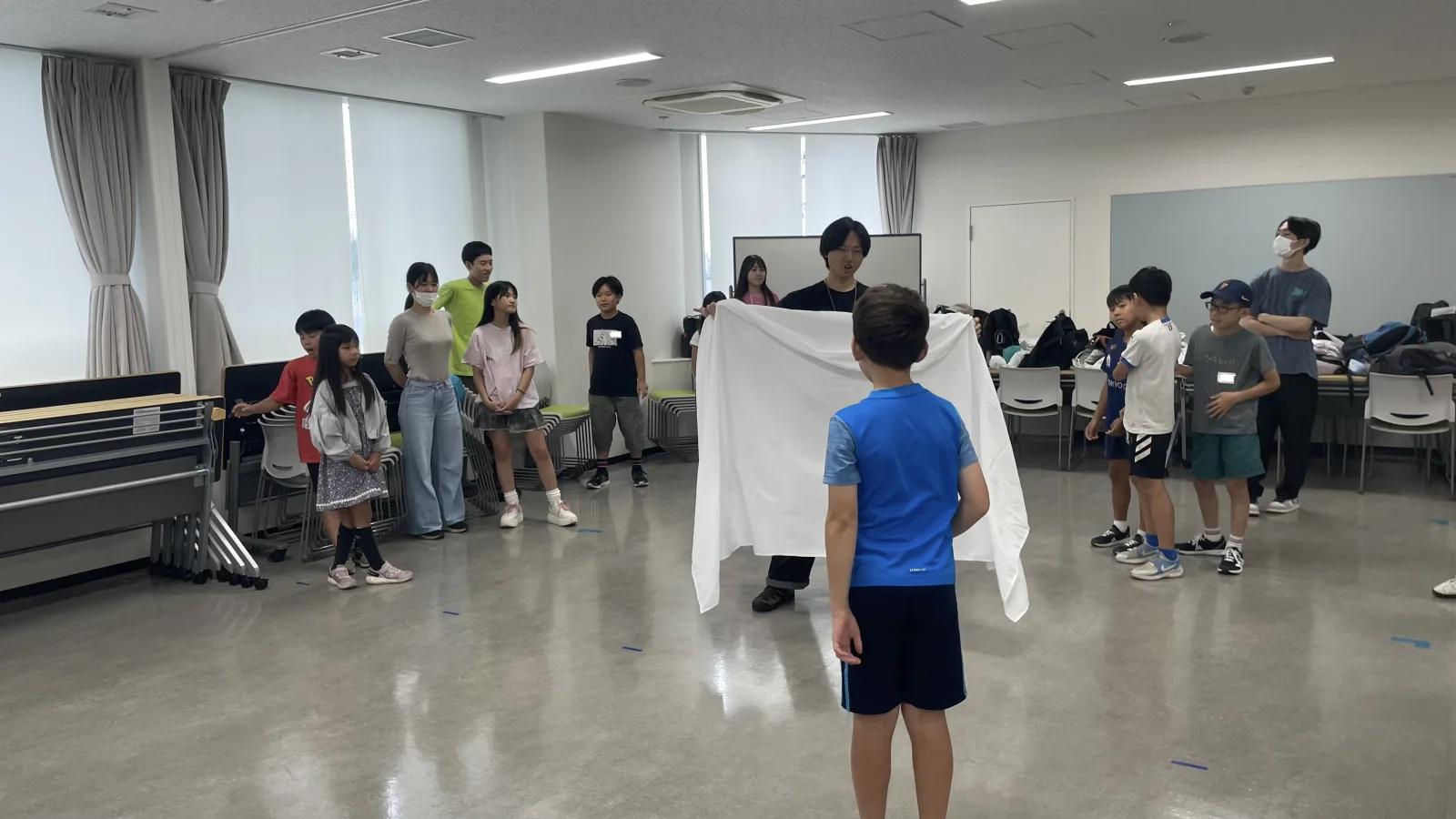 会議室。スタッフが白い布を広げて持ち、その手前に立つ子どもと、両側に分かれて見ている子どもたち