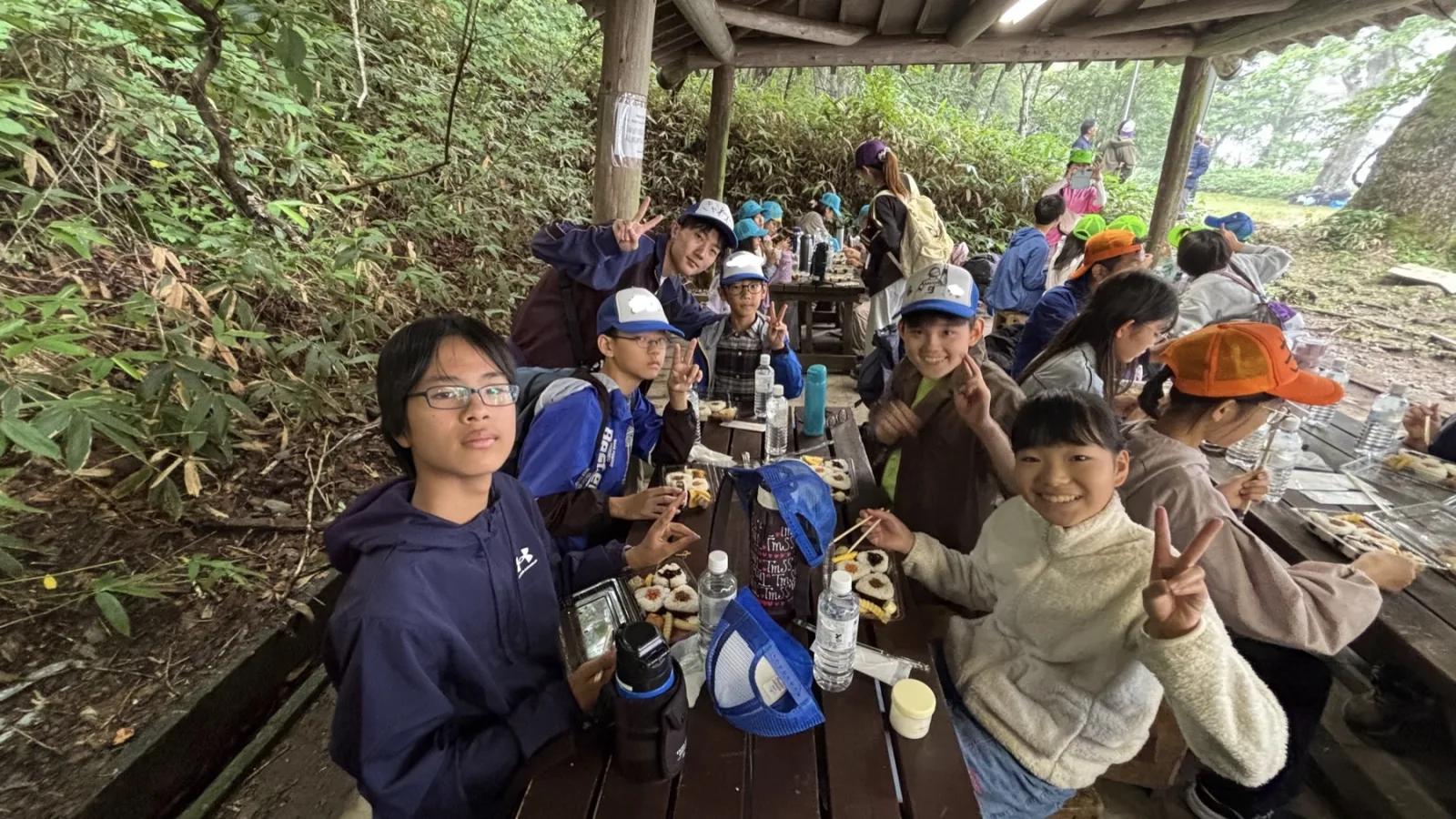 屋根付きの小屋で、長机を囲んだ参加者たちが箸を手にお弁当を食べながら、カメラに向かってピースをしている