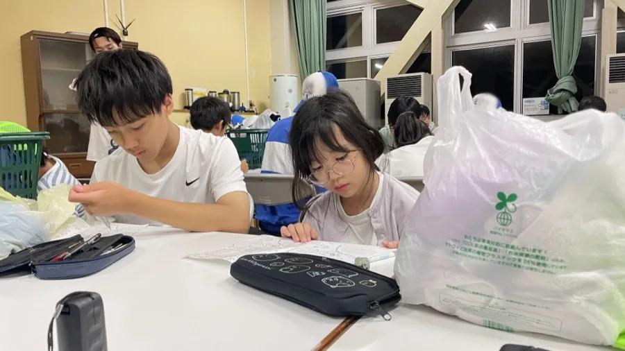 室内の白いテーブルで、しおりに書き込む参加者たち。窓の外はもう真っ暗
