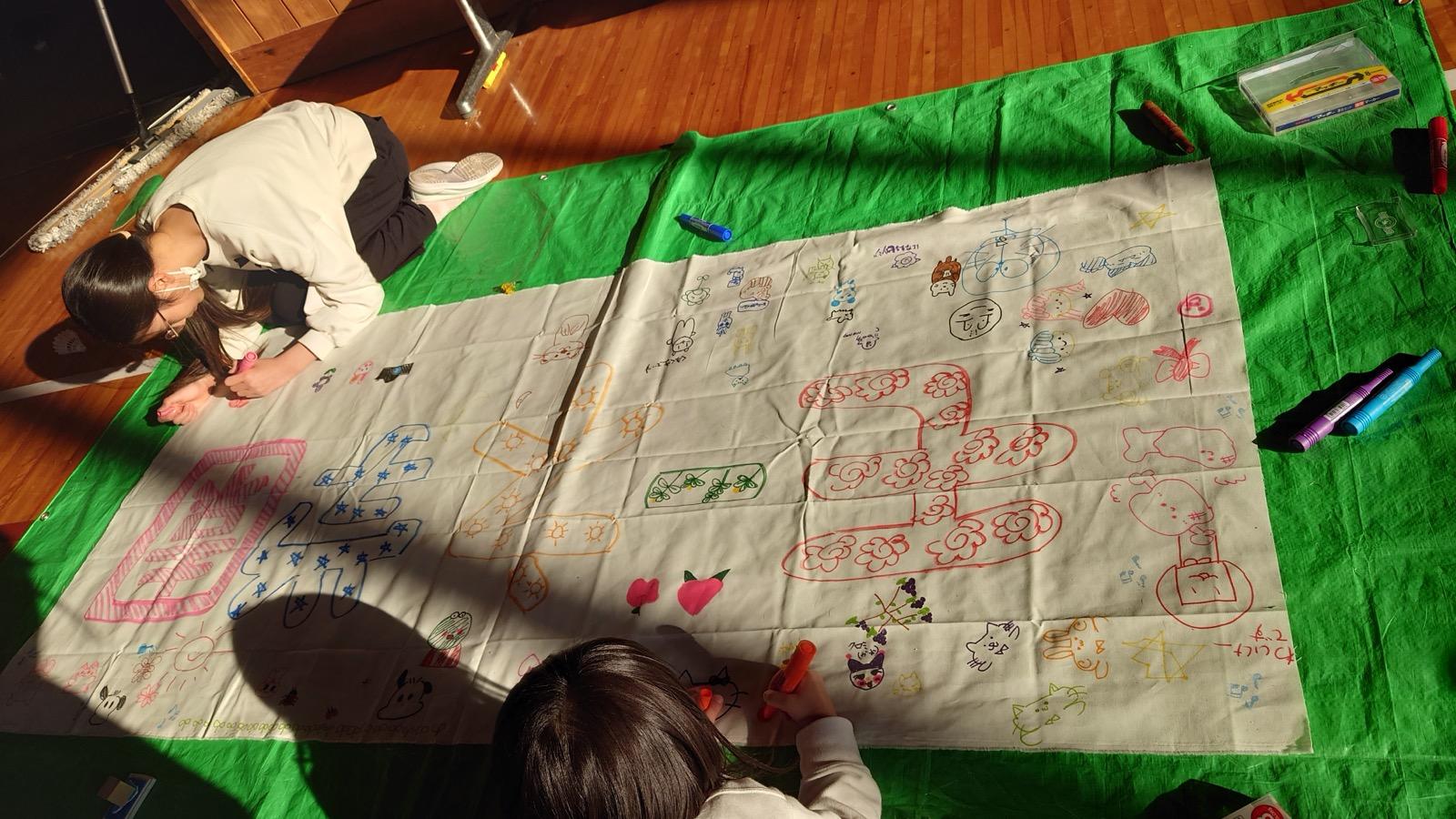 体育館の床に敷いた緑のシートの上で、大きな白い布にマジックで絵を描いている子どもたち