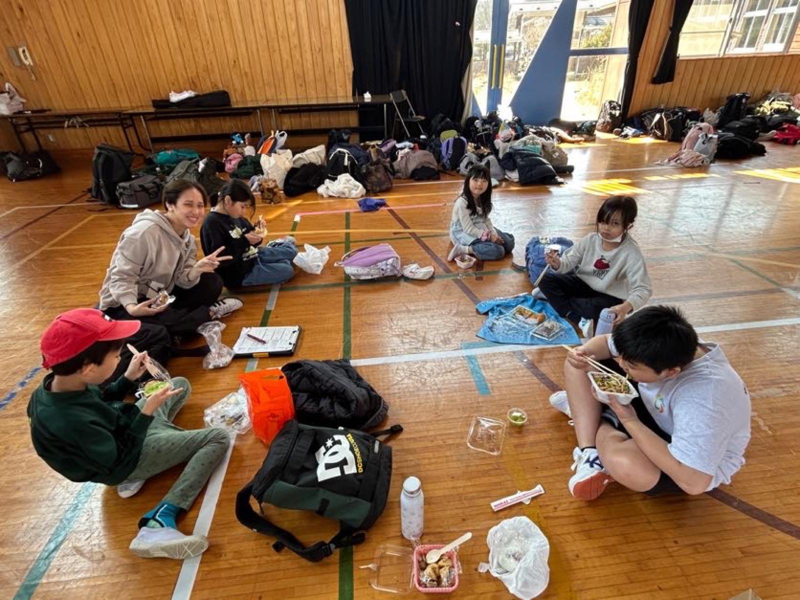 体育館の床に班ごとに固まって座り、お弁当を食べている子どもたち。壁際には荷物が並んでいる