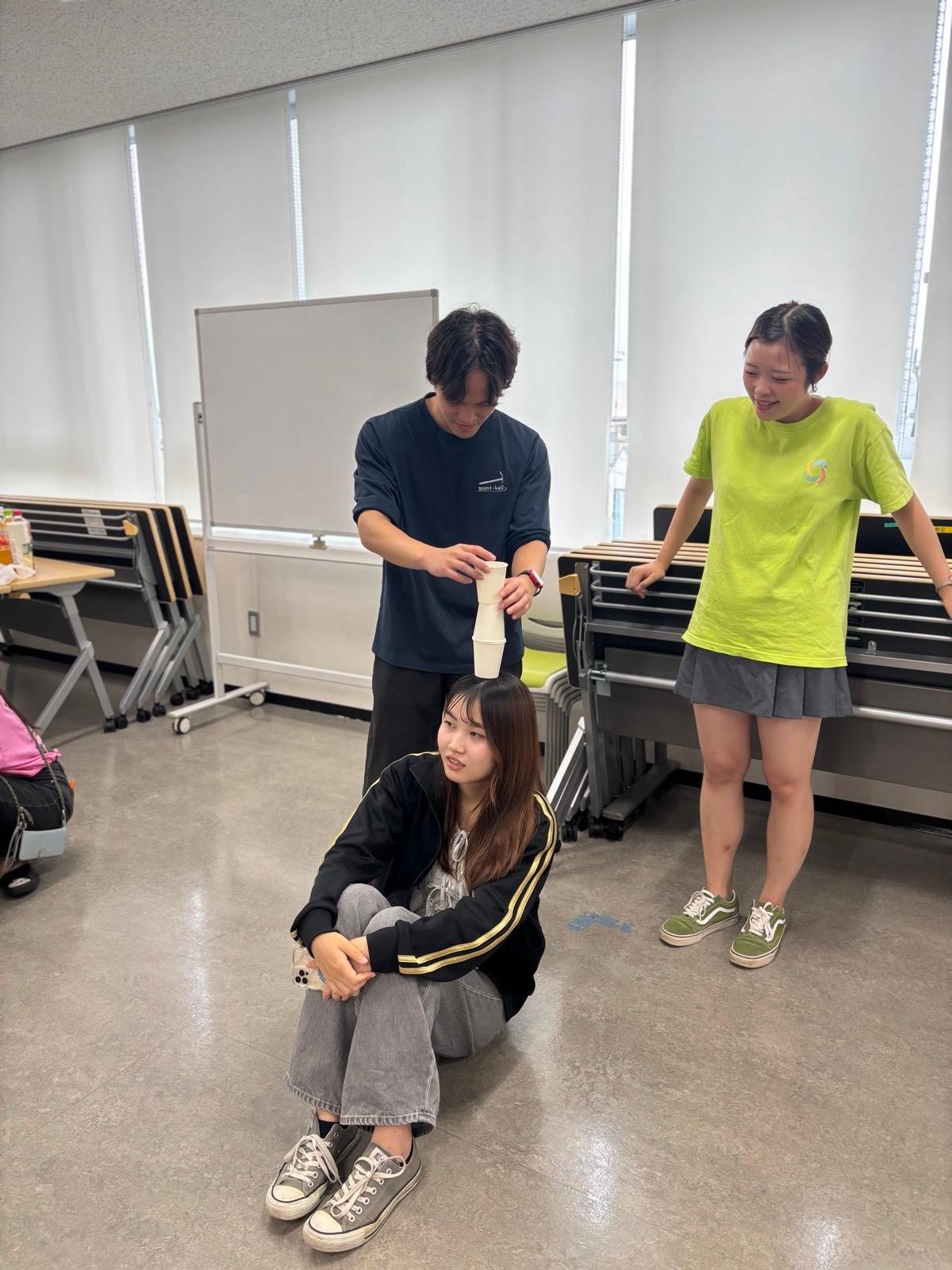 会議室で、床に座った人の頭の上にスタッフが紙コップを重ねて乗せようとしている。横で別のスタッフが笑っている