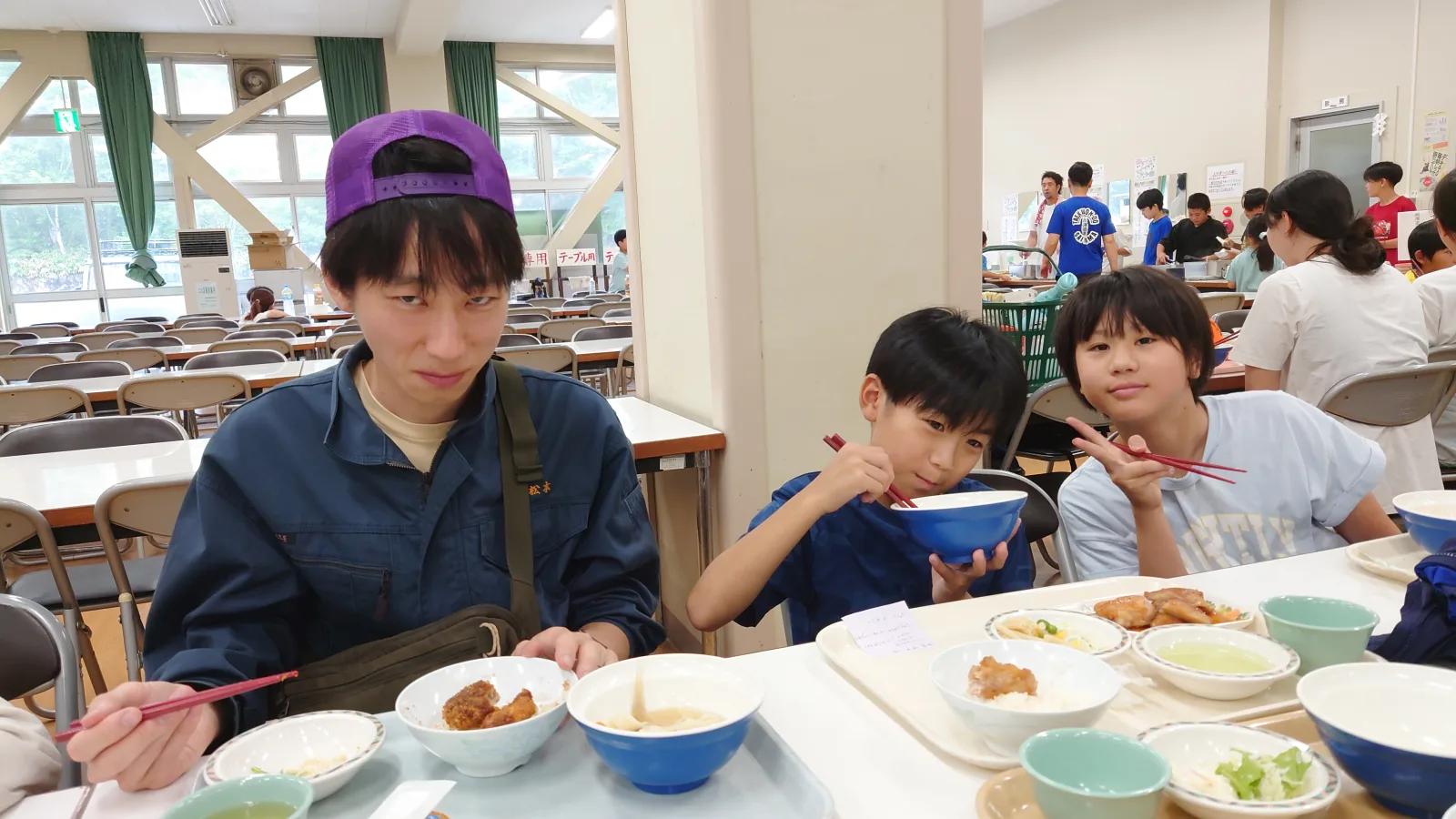 施設の食堂。長机で丼と小鉢の昼食を食べる参加者たちと、紫のキャップをかぶったスタッフ。奥では別のグループが配膳している