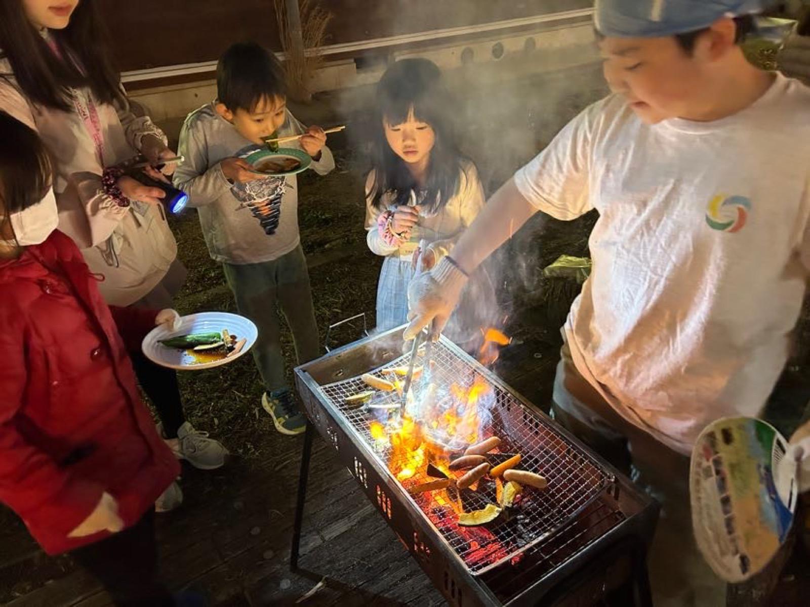 夜の中庭。バーベキューコンロの網の上でウインナーと野菜が焼かれ、軍手をした子どもがトングで返している。炎が上がっている