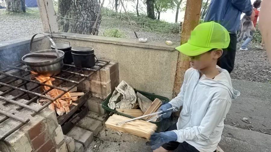 かまどで鍋を火にかけ、火ばさみと手袋を使って割った薪を追加している参加者