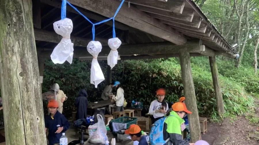 木造の東屋の梁から、白いビニール袋で作られたてるてる坊主が3つ、青いテープで吊り下げられている。奥では参加者たちが荷物を広げている