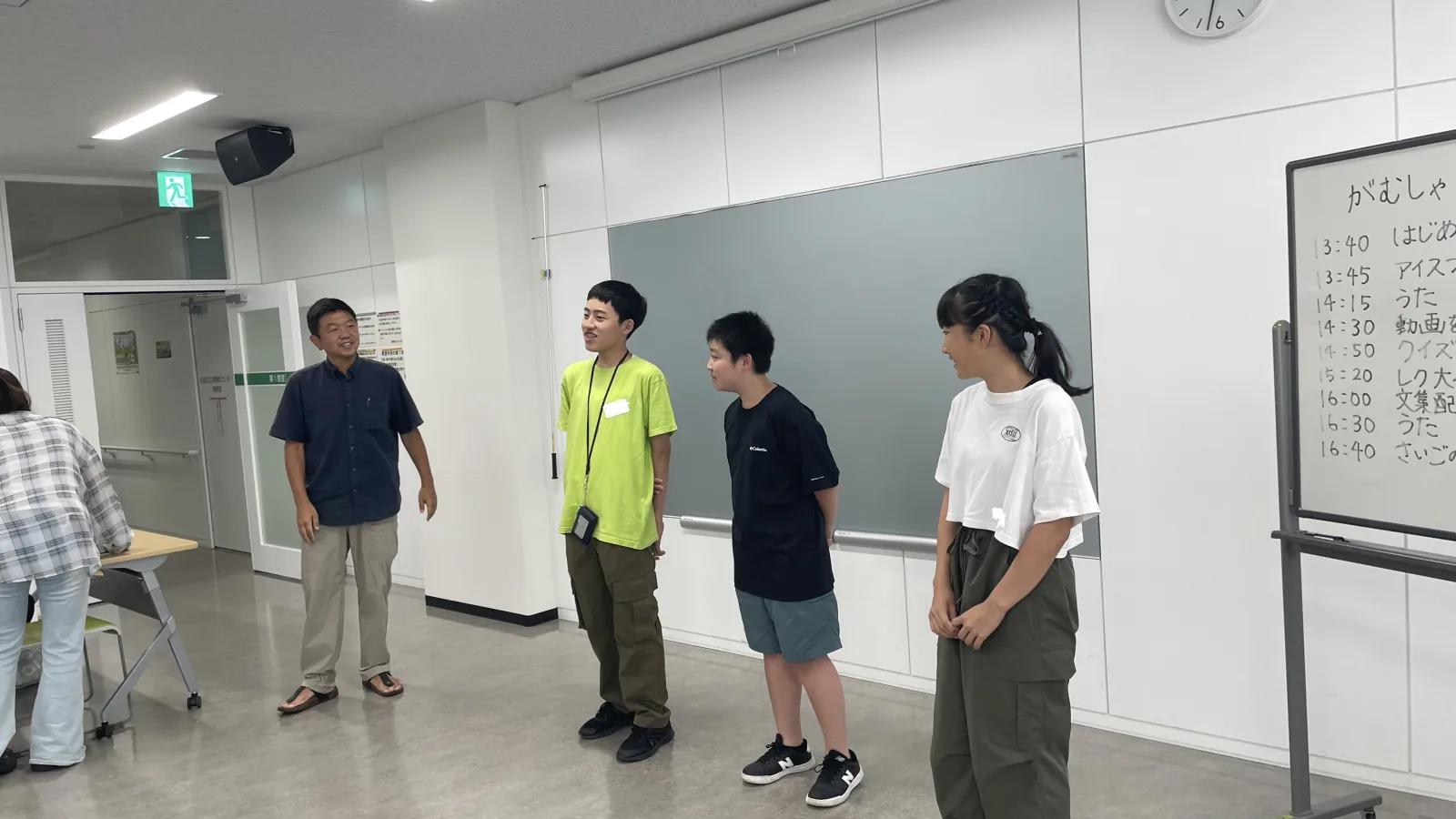 会議室。前に立った中学生3人とスタッフ。右手のホワイトボードには当日のタイムテーブルが手書きされている