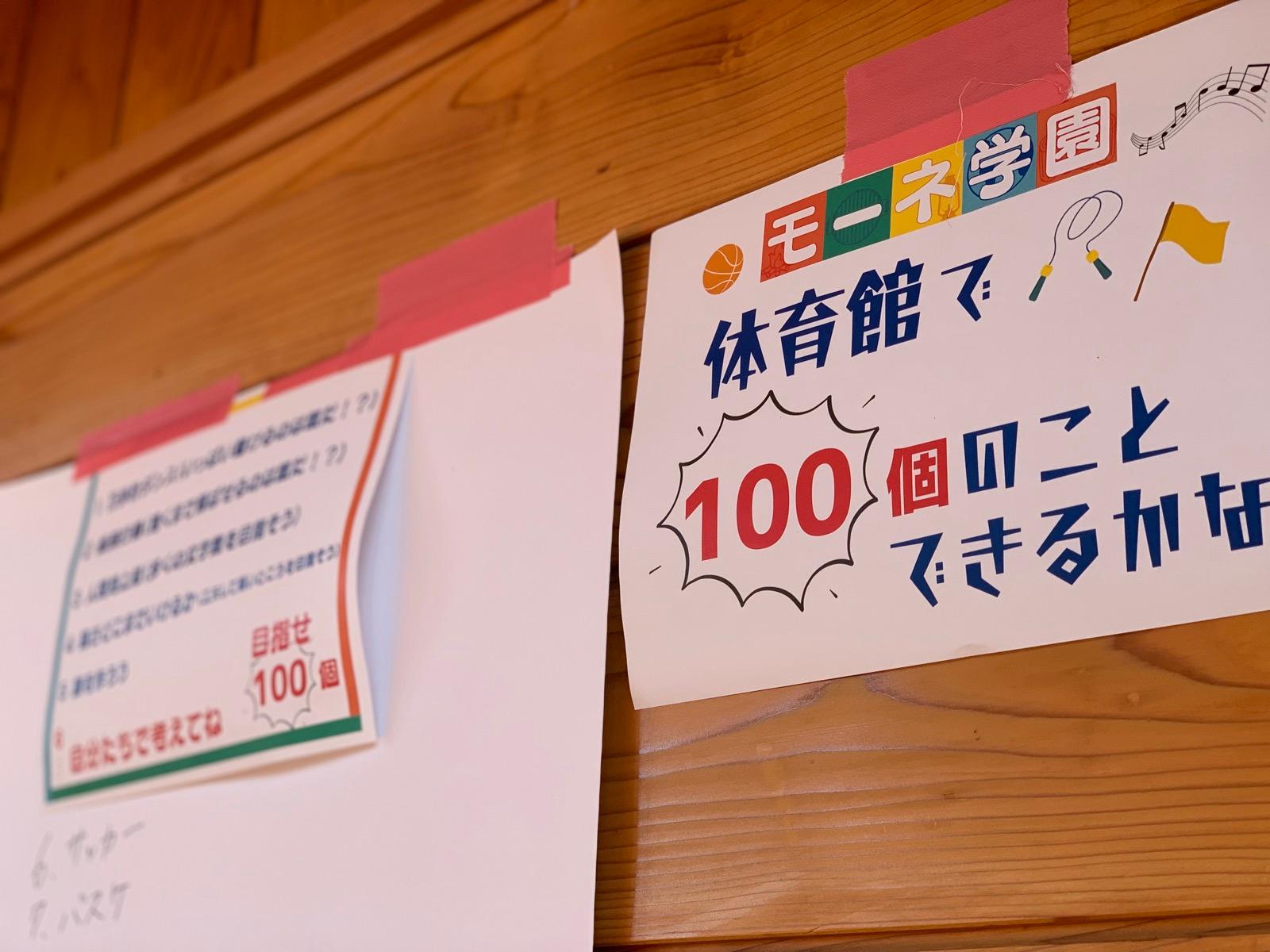 体育館の壁に貼られた模造紙。「モーネ学園 体育館で100個のことできるかな」と書かれ、ルールと項目が並んでいる。隣の紙には「6.サッカー」「7.バスケ」と追記されている