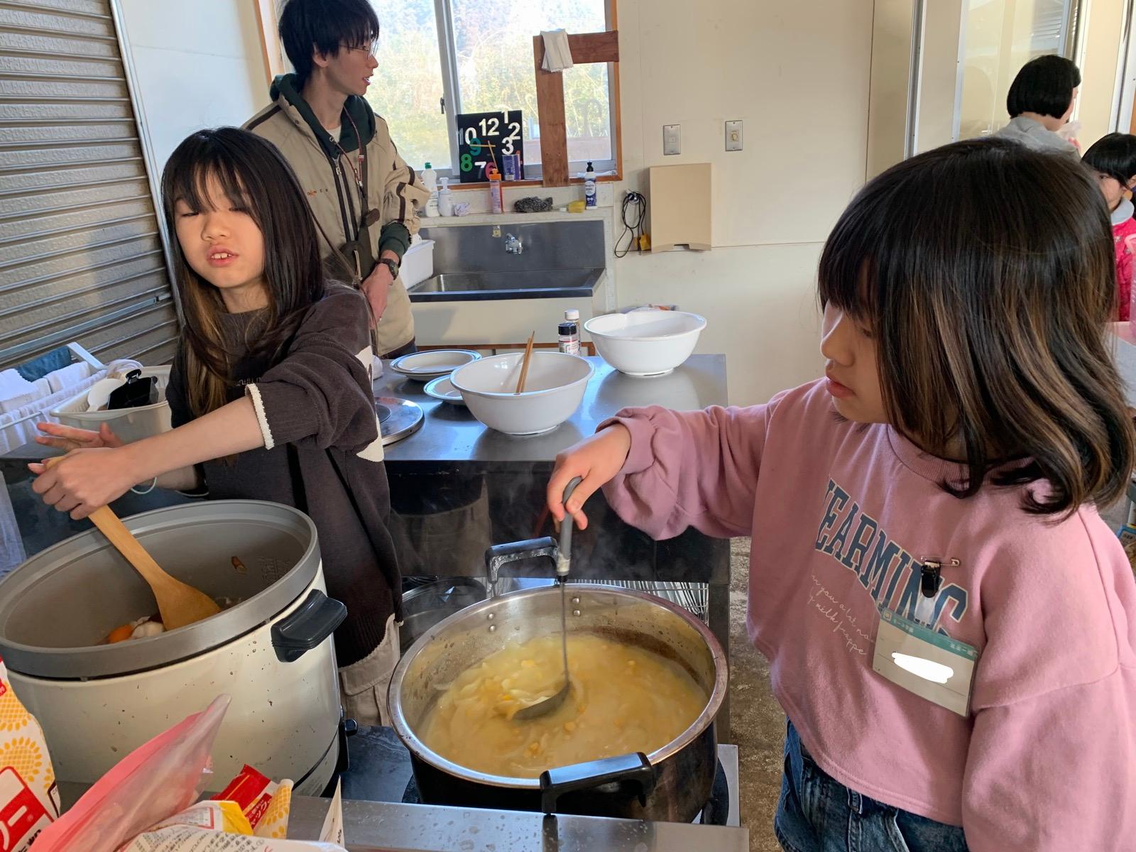 調理室で、二人の子どもが大きな寸胴鍋のスープをおたまでかき混ぜ、隣の炊飯器を木べらでかき混ぜている。湯気が上がっている