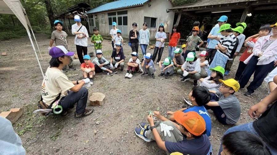 建物前の広場で車座になり、しゃがんだスタッフの話を聞く参加者たち。スタッフの手元には緑のテープと切り株、足元にはまきが置かれている