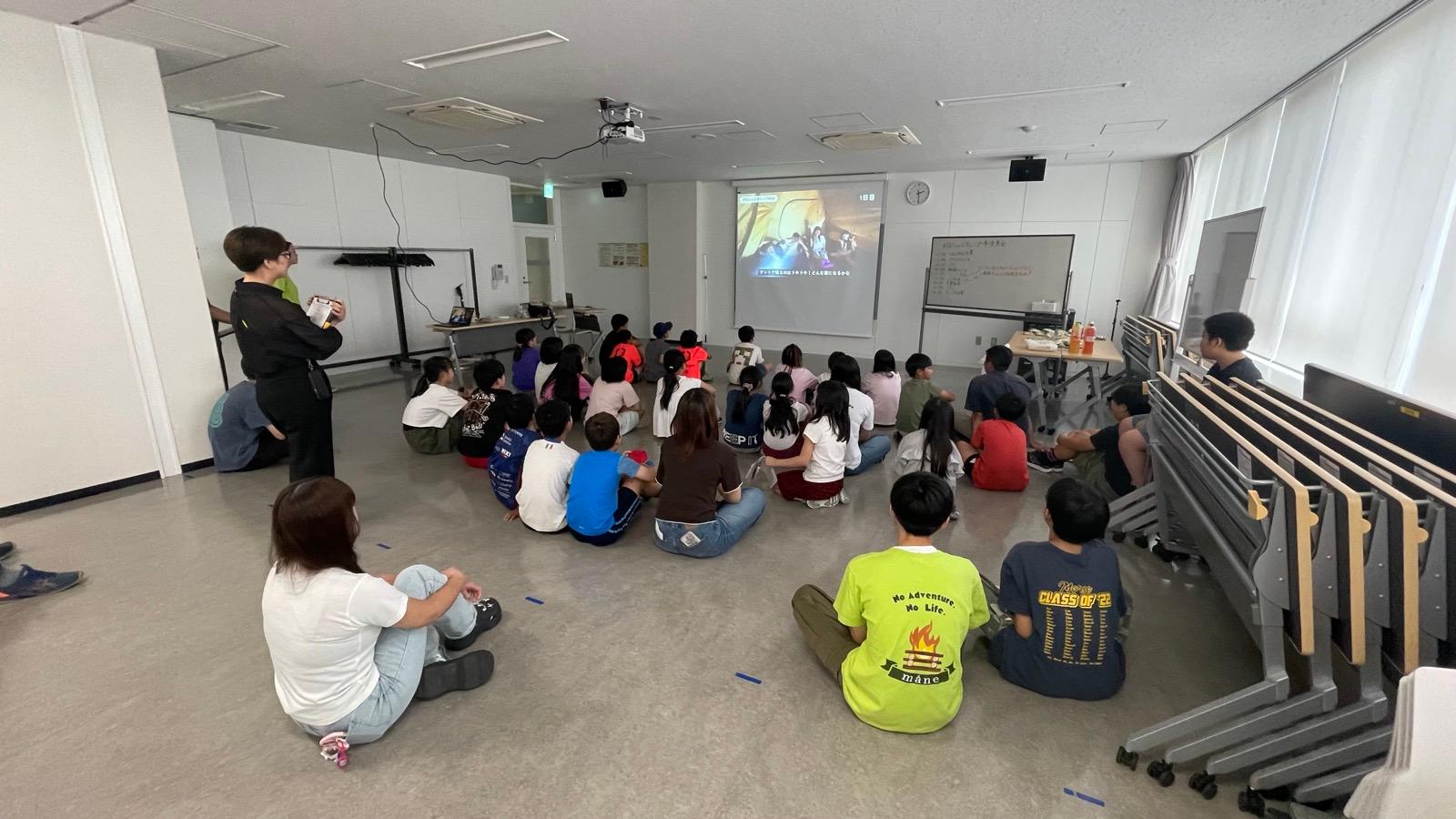 暗くした会議室で、プロジェクターの映像を床に座って見ている子どもたち。スクリーンにはテントの写真が映り、右上に「1日目」と出ている