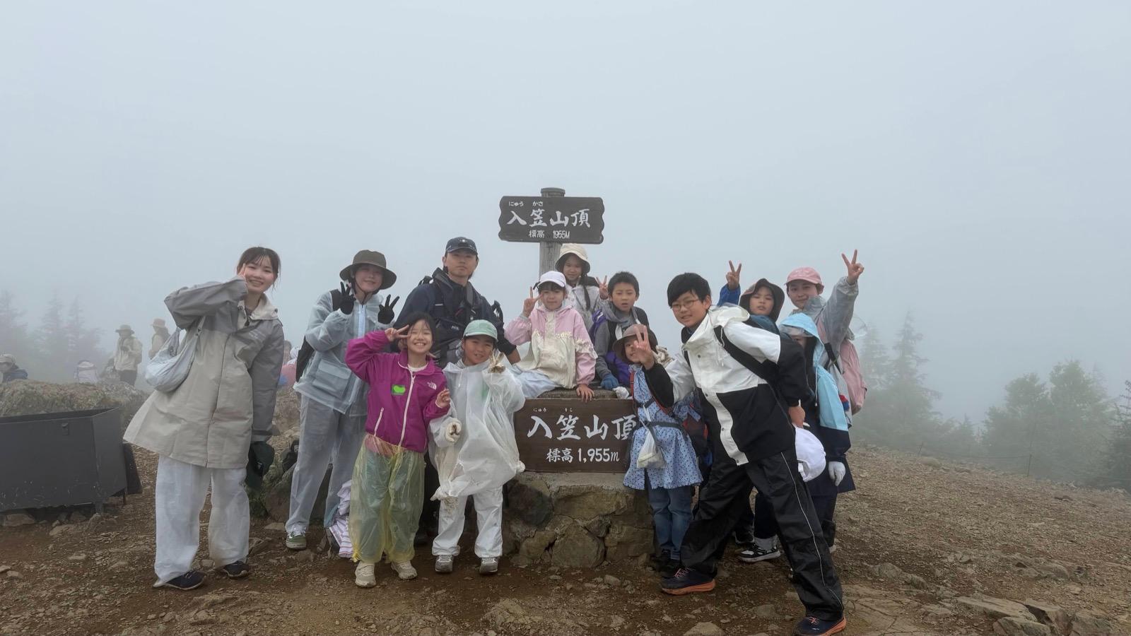 霧に包まれた山頂。「入笠山頂 標高1955M」の木の標識の周りに、レインウェアを着た子どもたちとリーダーが集まっている