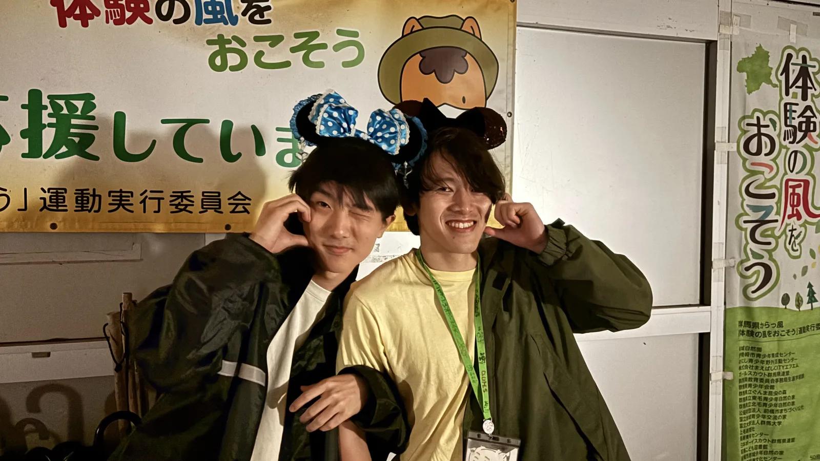 屋内で、頭にリボンのついた耳のカチューシャをつけたスタッフふたりが並んで笑っている。背景にはキャンプ関連の掲示物