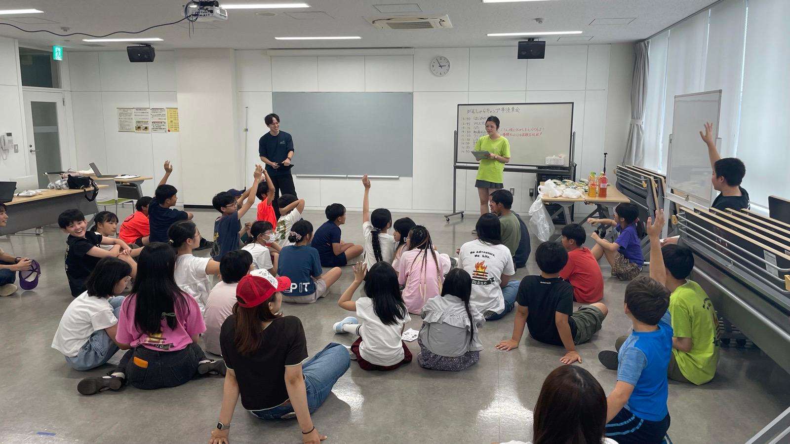 会議室。クリップボードを持ったスタッフが問題を読み上げ、床に座った子どもたちが手を挙げている。奥のホワイトボードには当日の予定、机にはお菓子とジュースが並ぶ