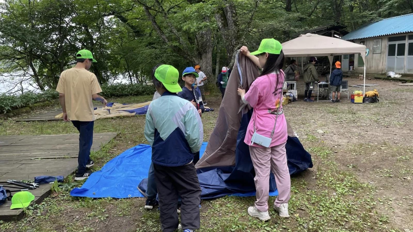 湖畔の広場で、黄緑色のキャップをかぶった参加者たちがブルーシートの上でテントの生地を広げてたたんでいる。奥にはタープと建物