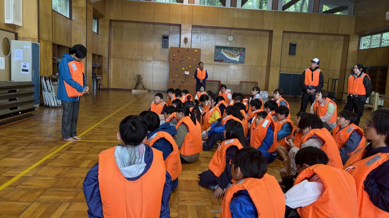 施設の広間。オレンジ色のライフジャケットを着た参加者が床いっぱいに座り、前に立ったスタッフの説明を聞いている