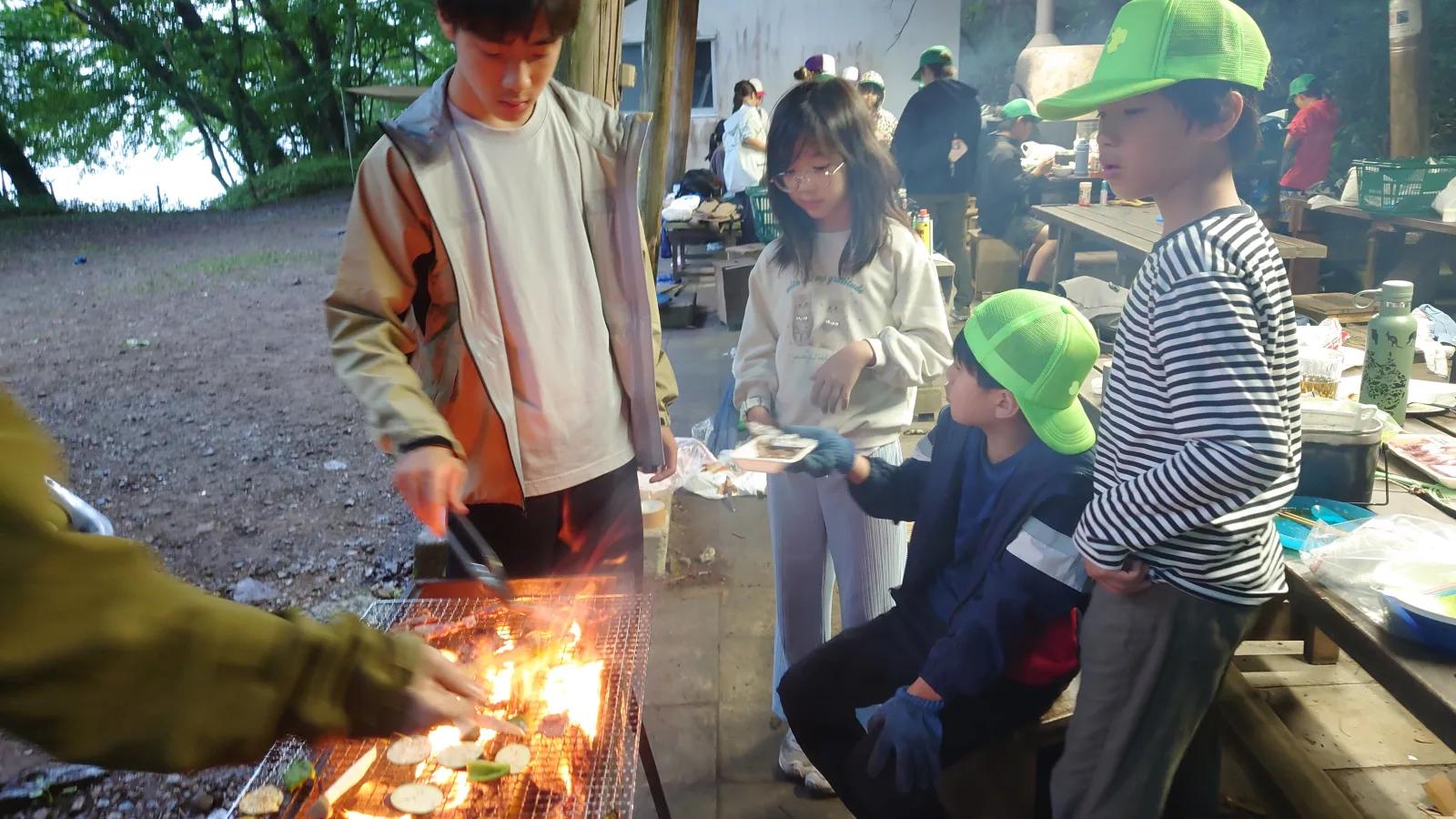 夕方の炊事場。金網のコンロで肉を焼くスタッフと、それを囲んで待つ参加者たち。奥では別の班も焼いている