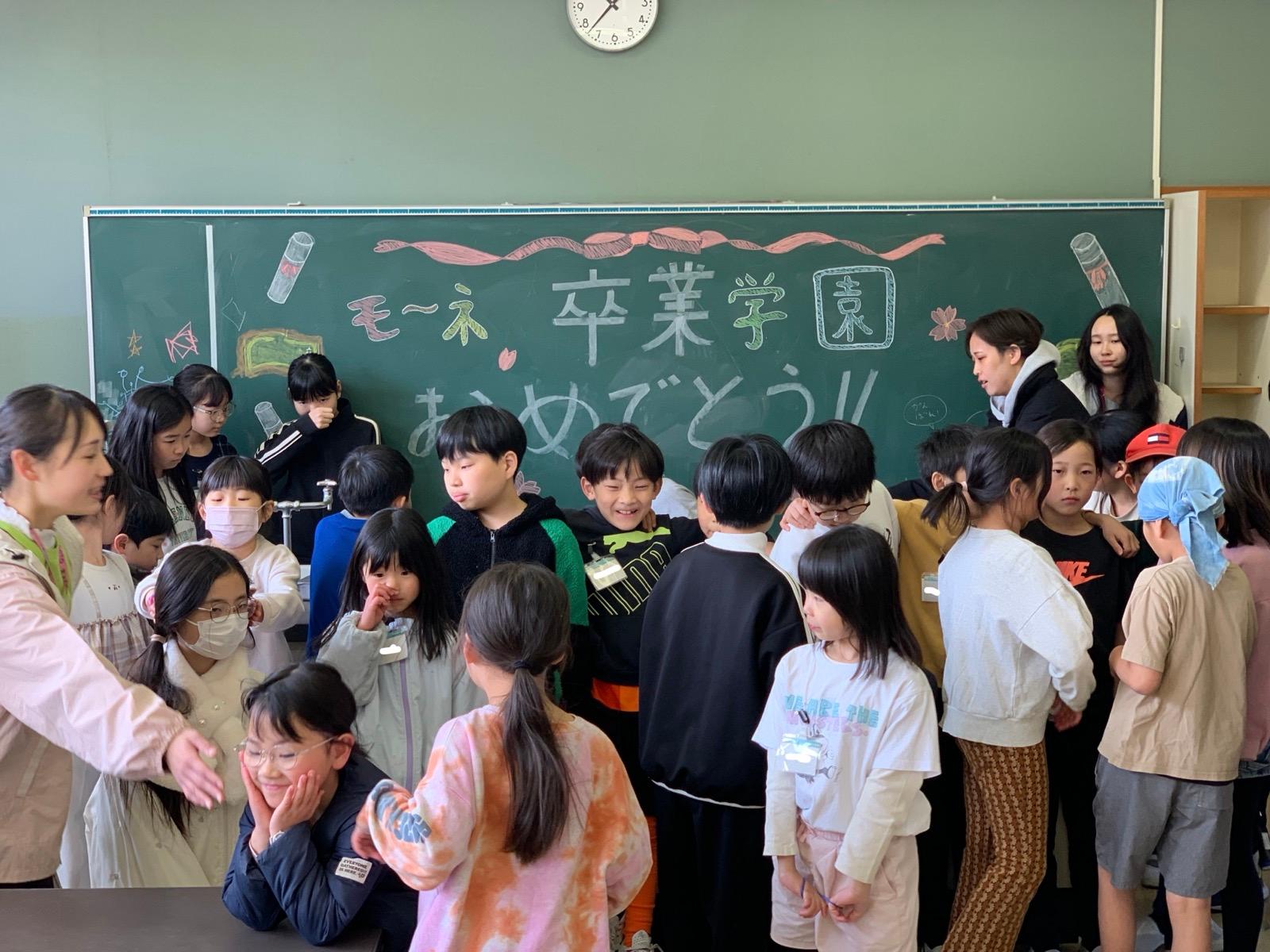 理科室の黒板の前に子どもたちが集まっている。黒板には「モ〜ネ 卒業学園 おめでとう!!」と書かれている
