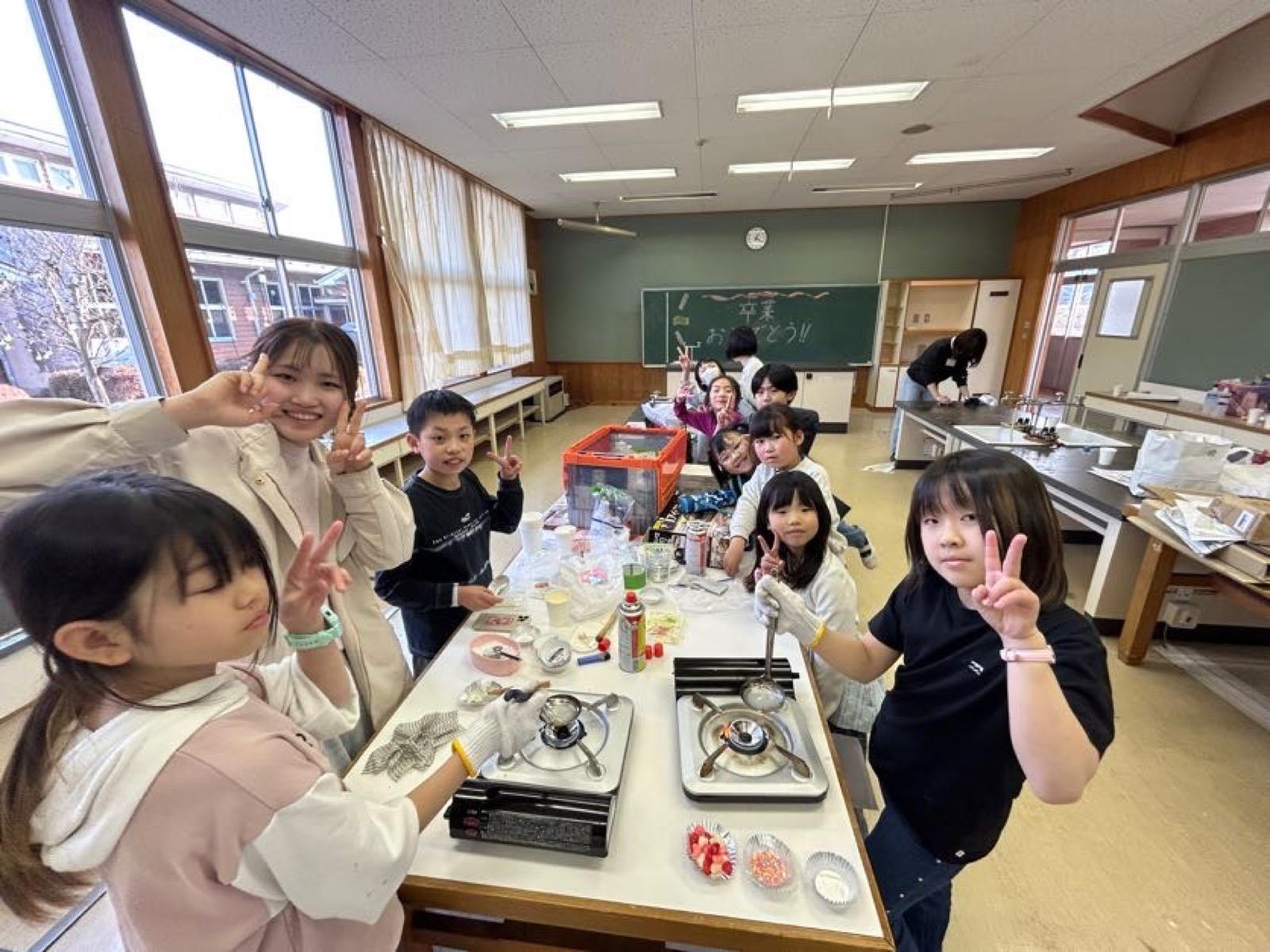 調理室。カセットコンロの上でお玉を火にかけ、子どもたちがのぞき込んでいる。手前のアルミカップには色とりどりのトッピングが入っている。黒板には「卒業おめでとう!!」と書かれている