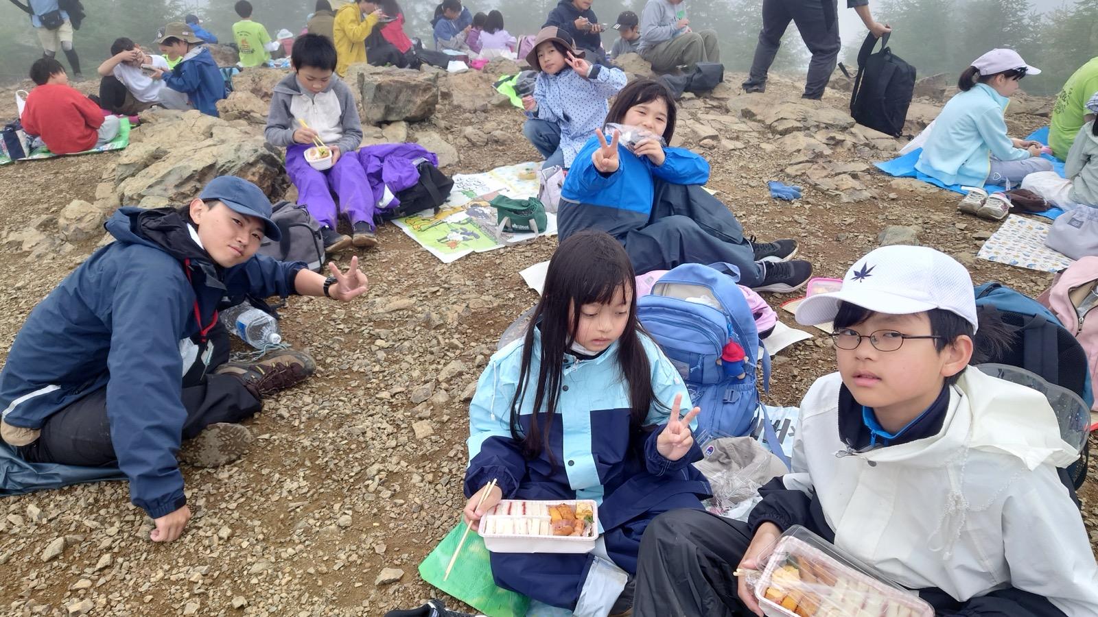岩がちの山頂の地面に座り込み、透明な容器の弁当を広げて食べている子どもたち。手前の子はサンドイッチと卵焼きの入った弁当を持つ。背景は一面の霧