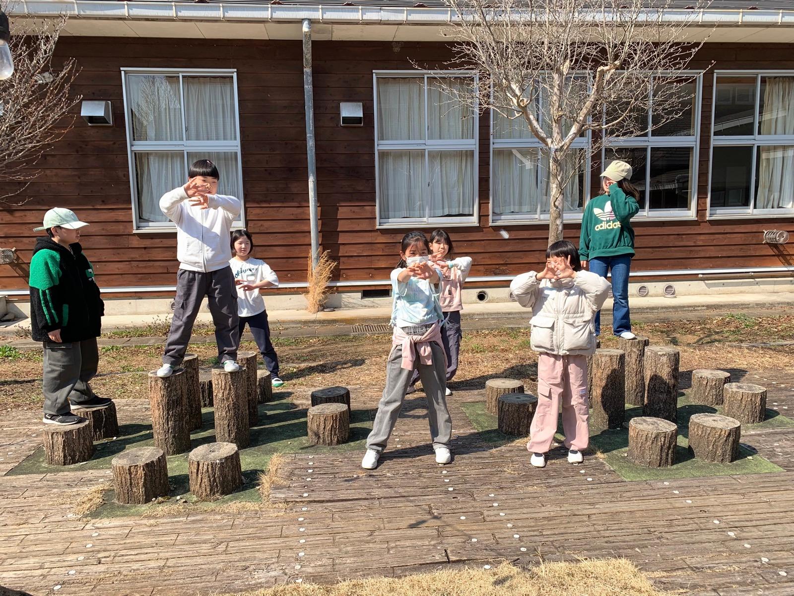 中庭に並んだ切り株の飛び石の上に、子どもたちが立って踊っている。木はまだ葉が出ていない