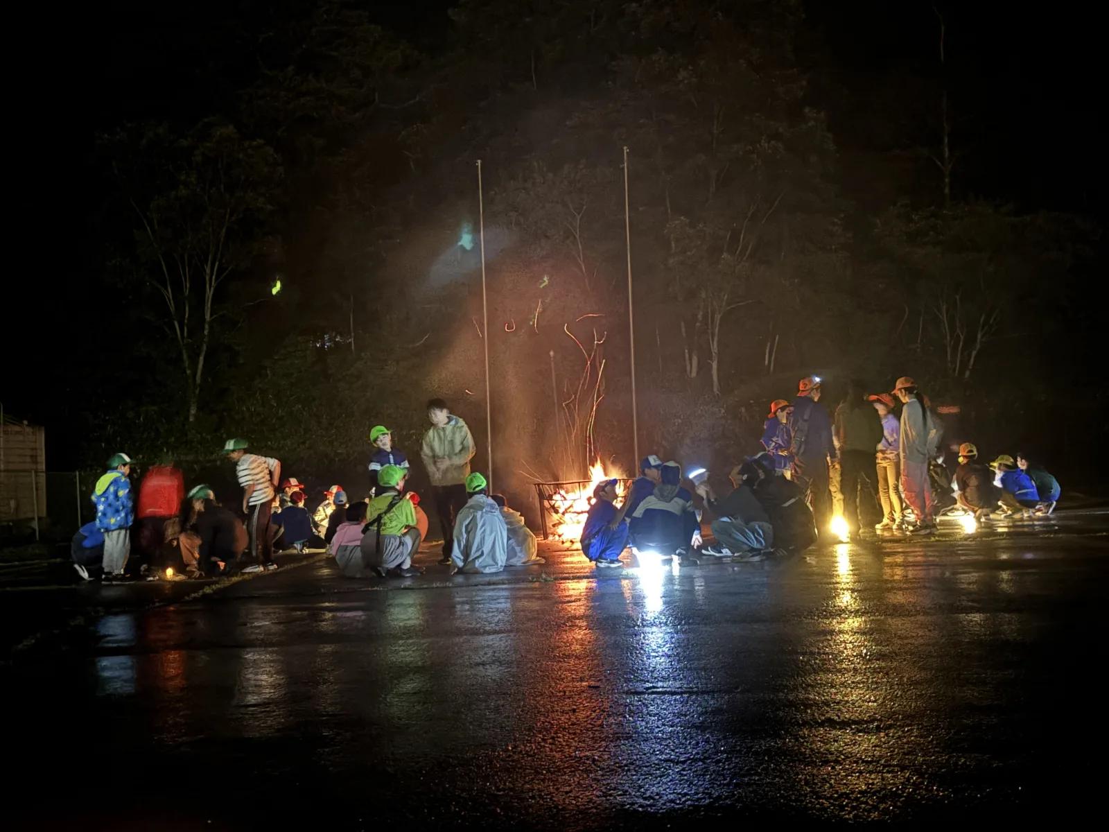 夜。雨に濡れたアスファルトの広場の中央で火が燃え、そのまわりを囲むように参加者たちが座ったり立ったりしている。地面に光が反射している