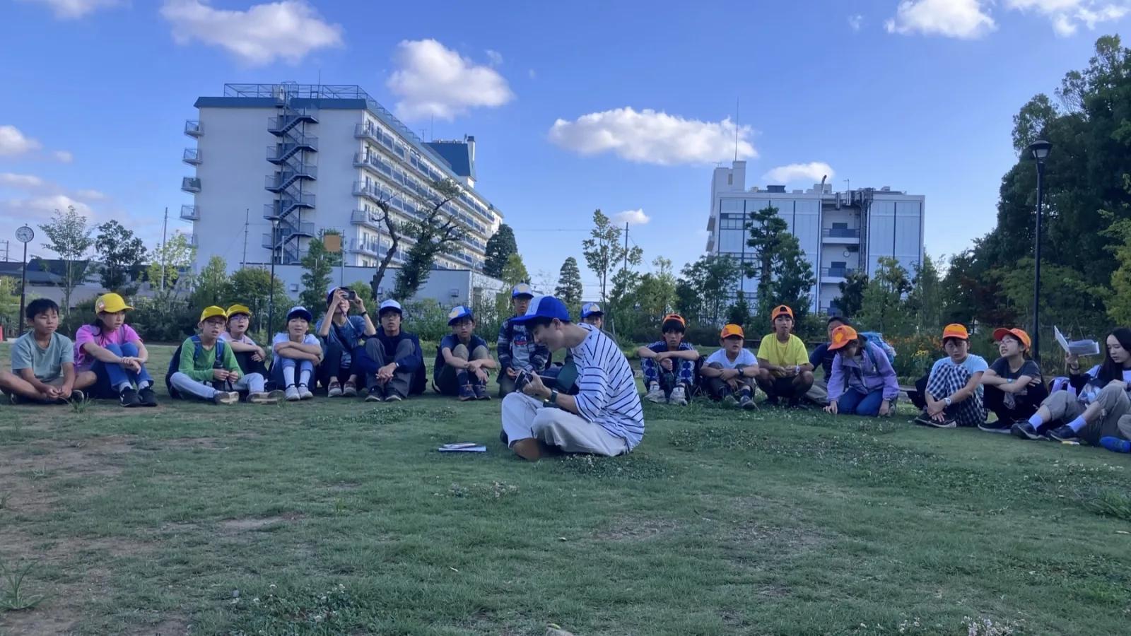 青空の下、芝生に横一列に座って前を見ている参加者たち。背景に建物と樹木
