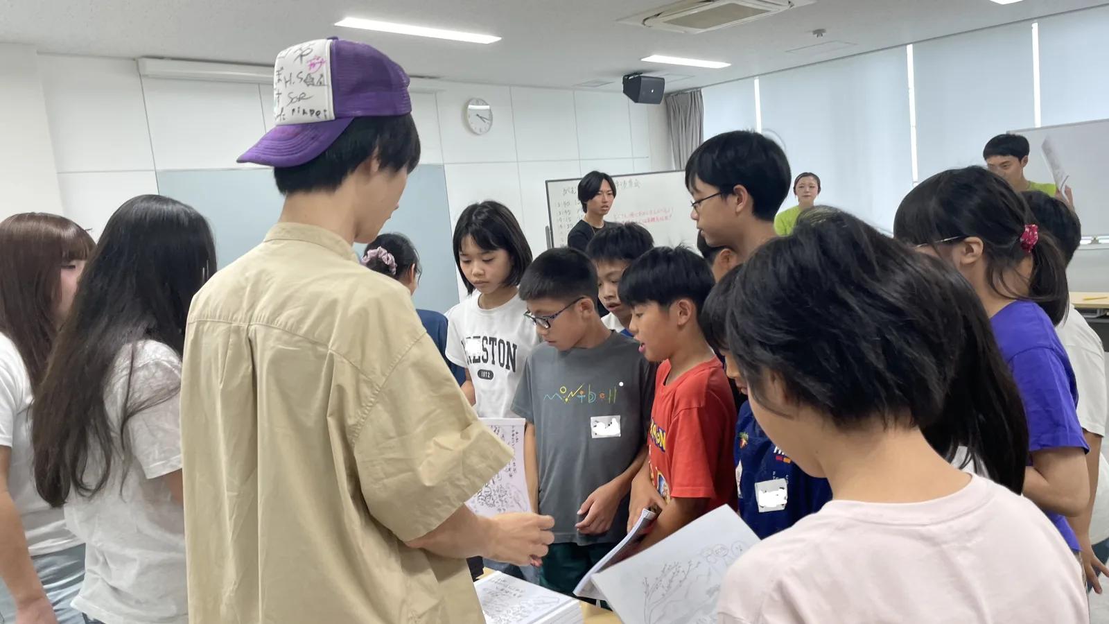 会議室で、キャップをかぶったスタッフの前に子どもたちが集まり、手描きの表紙の文集を受け取っている