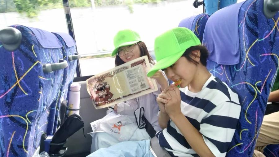 高速道路を走るバスの車内。百万円札の形をしたお菓子の袋を掲げて笑う参加者と、隣でグミをかじる参加者