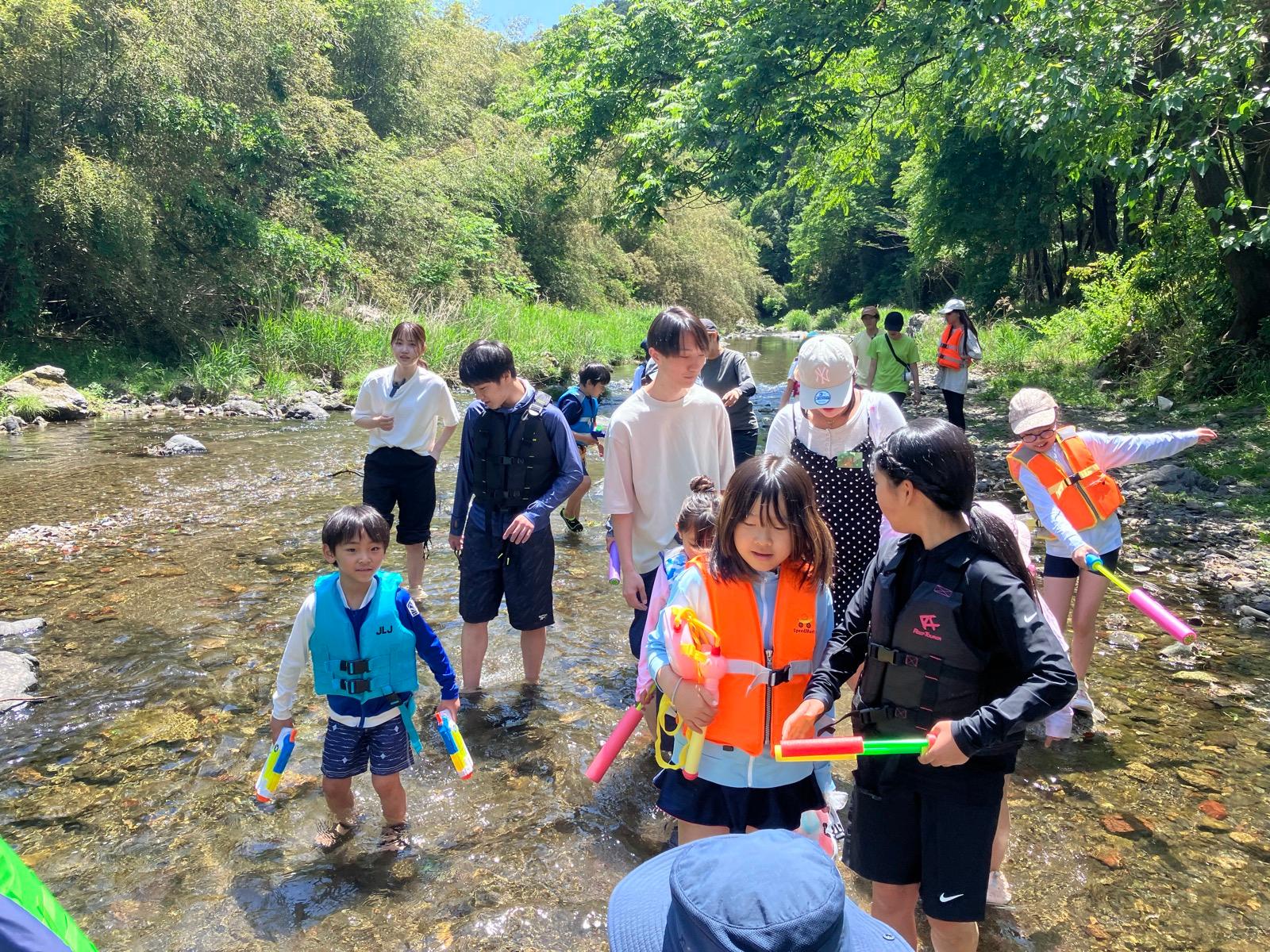 浅瀬に立ち、オレンジや青のライフジャケットを着て水鉄砲を構える子どもたち